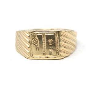 May include: A gold signet ring with a rectangular face. The face features an engraved design with the letters "HR" and a textured background. The ring has a ribbed design on the sides.
