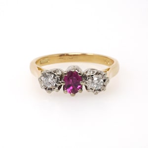 May include: A gold ring featuring a central oval-cut vibrant pink gemstone flanked by two round-cut clear white diamonds. The stones are set in a white metal setting that contrasts with the polished yellow gold band of the ring.