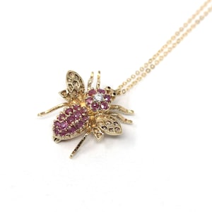 Puede incluir: Un collar con colgante de abeja dorado. El cuerpo de la abeja está adornado con pequeñas piedras preciosas rosas, y las alas tienen detalles intrincados. Un solo diamante está engastado en el centro de la cabeza de la abeja. La cadena del collar también es dorada.