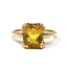 May include: A gold ring featuring a large, rectangular yellow gemstone. The gemstone is the focal point, set in a gold band with decorative accents. The ring is displayed against a white background.