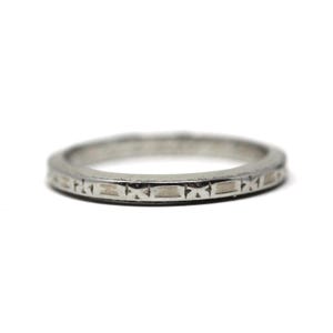May include: A silver ring with a repeating rectangular and starburst pattern around the band. The ring is set against a white background, highlighting its intricate design and metallic sheen. This piece is a classic jewelry item.