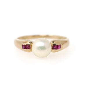 May include: A gold ring featuring a central round white pearl flanked by two small rectangular red rubies on each side. The band is smooth and polished, showcasing a classic design with a prominent pearl setting and vibrant gemstone accents.