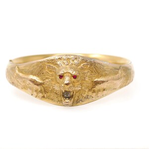 May include: A gold bangle bracelet featuring a central relief of a roaring lion head. The lion has two small red gemstone eyes and a single clear gemstone set within its open mouth. The metal surface displays detailed engraved fur textures.