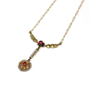Puede incluir: Un collar vintage dorado con un colgante central con una piedra preciosa roja rodeada de pequeñas perlas. El collar tiene una cadena delicada y detalles en forma de hojas, mostrando un diseño antiguo.
