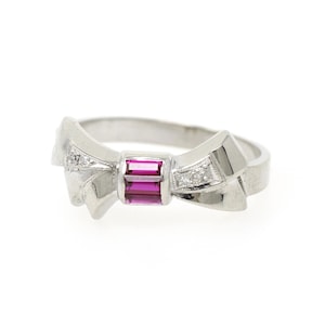 May include: A silver-toned ring featuring a bow-shaped centerpiece. The center of the bow is set with a rectangular, vibrant pink gemstone, while the sides of the bow are accented with small, clear, sparkling stones embedded in the metal.