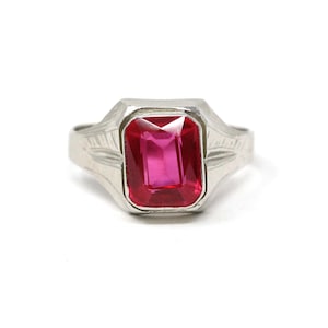 Puede incluir: Un anillo plateado con una gran gema rectangular de color rosa brillante. El anillo tiene un diseño grabado en la banda. La gema está engastada en un bisel plateado, sobre un fondo blanco.