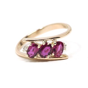 May include: A gold bypass ring featuring three oval-cut vibrant pink gemstones set in a row, flanked by two small clear accent stones. The band has a smooth, polished finish and a curved, open design that holds the stones between two parallel bars.