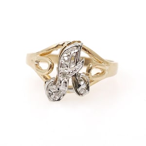 May include: A gold ring featuring a central letter A in a silver-toned metal finish. The letter is embellished with small, clear, sparkling stones. The ring band is crafted from yellow gold and displays an open, decorative, swirling design.