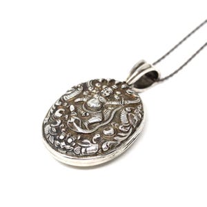 May include: Oval silver pendant with intricate relief carvings. The pendant features a detailed scene with figures, birds, and floral elements. It hangs from a delicate silver chain, creating a classic jewelry piece.