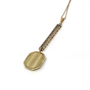 May include: A gold-coloured pendant necklace with an Art Deco design. The necklace features a rectangular bar with geometric cutouts and a textured, octagonal pendant. The pendant has a striped pattern and hangs from a delicate chain.