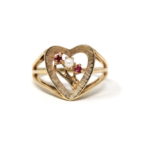 Puede incluir: Anillo dorado con diseño en forma de corazón y superficie texturizada. El corazón enmarca una pequeña rama con una perla y dos gemas rojas. El anillo tiene un diseño de doble banda.
