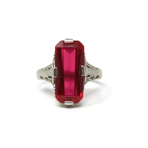 Könnte beinhalten: Ein silberner Ring mit einem großen, rechteckigen, rubinroten Edelstein. Der Ring hat ein verziertes Filigranmuster auf dem Band und um die Edelsteinfassung. Der Edelstein ist der Mittelpunkt, mit seiner leuchtenden Farbe und Form.