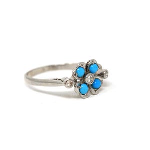 K&ouml;nnte beinhalten: Silberring mit einem Blumenmuster, besetzt mit f&uuml;nf leuchtend blauen T&uuml;rkissteinen und einem zentralen, klaren Edelstein. Der Ring hat ein zartes Band mit kleinen Akzentsteinen auf beiden Seiten der Blume.