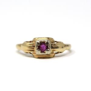 Puede incluir: Un anillo de oro con una piedra preciosa roja oscura cuadrada engastada en bisel. El anillo tiene un diseño detallado y texturizado en la banda y alrededor del engaste de la piedra preciosa. La piedra preciosa es de un color vibrante y llamativo.