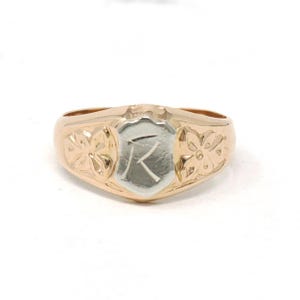 May include: A gold ring featuring a central shield-shaped plaque with a silver-toned finish and the engraved letter K. The band is crafted from polished gold and displays embossed floral motifs on both sides of the central shield element.