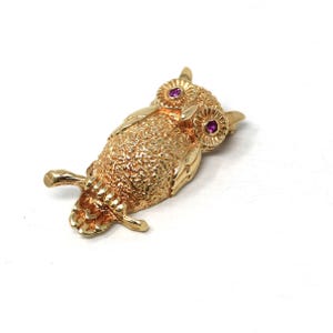 Op de afbeelding: Een goudkleurige uilbroche met getextureerde details. De ogen van de uil zijn klein, rond en voorzien van een dieproze edelsteen. De broche is een gedetailleerde weergave van een uil, met vleugels en poten.