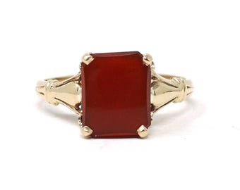 May include: A gold ring with a rectangular, deep red gemstone. The ring has a classic design with decorative elements on the band and around the stone. The gemstone is the focal point, showcasing a rich, warm colour.
