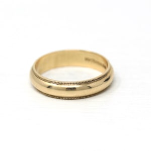 May include: A gold wedding band with a rounded profile and a detailed beaded edge. The ring is made of polished gold and sits on a white surface. The band is a classic design.