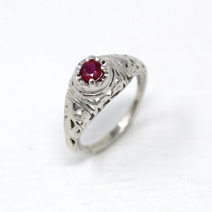 Puede incluir: Un anillo de plata con una gema redonda de color rojo intenso. El anillo tiene un diseño de filigrana ornamentado con patrones calados. La gema está engastada en un bisel elevado, y la banda del anillo es lisa y pulida.