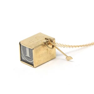 Puede incluir: Collar con colgante dorado con un dije en forma de cubo. El dije muestra un billete de dólar y el texto "IN EMERGENCY BREAK GLASS". Un pequeño dije de flecha dorada está adjunto.