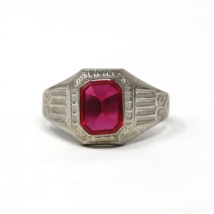 Puede incluir: Un anillo plateado con una gema rectangular de color rosa intenso en el centro. El anillo tiene un diseño detallado y texturizado alrededor de la gema y en la banda, de estilo antiguo o vintage.