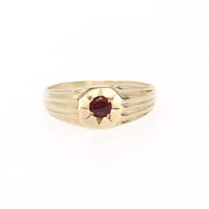May include: A gold ring featuring a central round dark red gemstone set within a starburst engraving. The band has a polished finish with horizontal grooved detailing extending from the centre setting towards the sides of the ring.