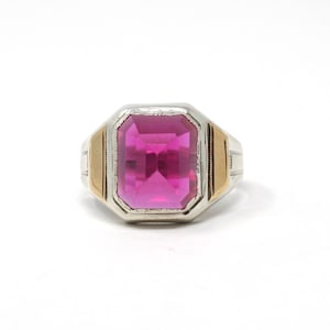May include: A silver ring with a large, rectangular, bright pink gemstone. The ring has gold accents on the sides and a detailed design on the band. The gemstone is the focal point, catching the light and adding a pop of color.