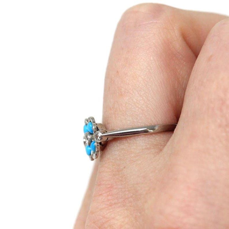 K&ouml;nnte beinhalten: Ein silberner Ring mit einem bl&uuml;tenf&ouml;rmigen Design, besetzt mit sechs hellblauen T&uuml;rkissteinen und kleinen klaren Steinen. Der Ring wird an einem Finger gezeigt, wobei der Fokus auf den Details und dem Farbkontrast liegt.