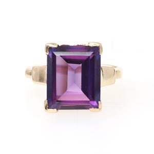 May include: A gold ring featuring a large, rectangular, emerald-cut purple amethyst gemstone set in a four-prong mounting. The band is made of polished yellow gold and connects to the setting with a simple, clean design that highlights the stone.
