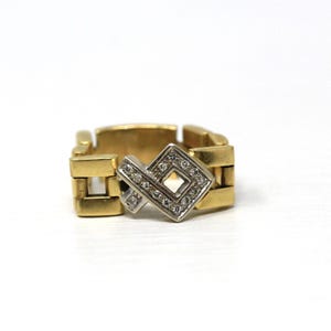 May include: A gold-coloured ring with a geometric design. The ring has a series of rectangular links forming the band. A silver-coloured, diamond-studded square and angled design is centered on the ring, adding sparkle.