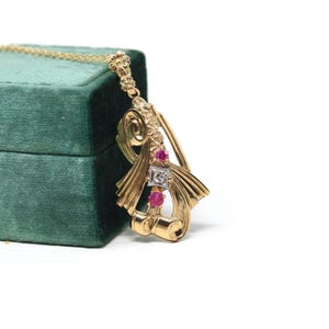 Puede incluir: Un collar con colgante de oro con un diseño floral en la parte superior. El colgante presenta un diamante cuadrado y dos piedras preciosas rojas. El collar se exhibe sobre una superficie de terciopelo verde.