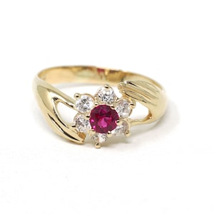 May include: A gold ring featuring a central round-cut vibrant red gemstone surrounded by six clear, round-cut white stones arranged in a floral pattern. The band is crafted from polished yellow gold and features two stylized hands cradling the stone cluster.