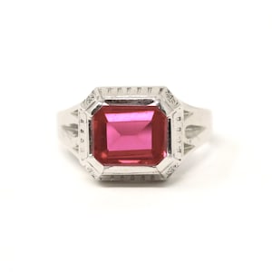 Puede incluir: Un anillo de plata con una gran gema rosa brillante de forma rectangular. La gema está engastada en un bisel decorativo y geométrico. El anillo tiene un diseño clásico y elegante.