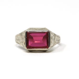 May include: A silver-toned ring with a large, rectangular, ruby-red gemstone. The ring has an engraved floral design on the band and around the gemstone setting. The gemstone is the focal point, showcasing a vibrant, deep red hue.