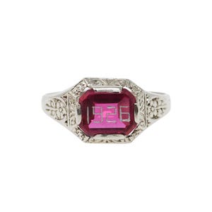May include: A silver-toned ring featuring a rectangular, deep pink gemstone set in a decorative metal frame. The center of the stone is engraved with the number 1926. The band displays intricate floral and scrollwork patterns along the sides.