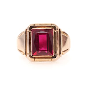 May include: A gold ring featuring a large, rectangular, faceted deep red gemstone set in a structured, polished gold band. The setting has a geometric, stepped design surrounding the central stone, creating a bold and symmetrical appearance.