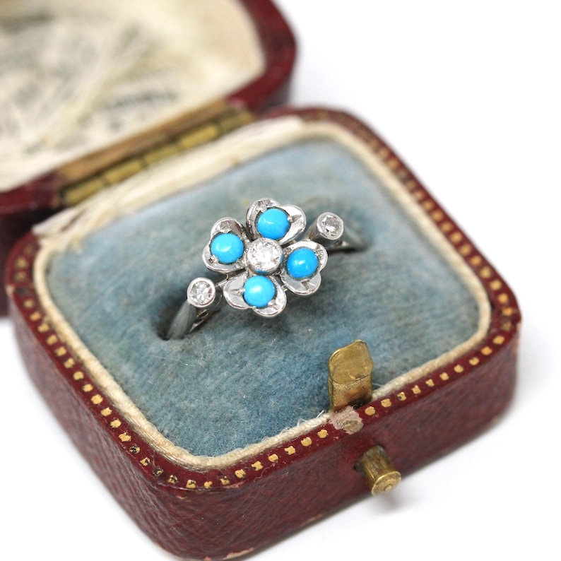 K&ouml;nnte beinhalten: Ein silberner Ring mit Blumenmuster, besetzt mit T&uuml;rkissteinen und einem zentralen Diamanten. Der Ring wird in einer roten Vintage-Schmuckschatulle mit blauem Samt pr&auml;sentiert, die die Details und Farben des Rings hervorhebt.