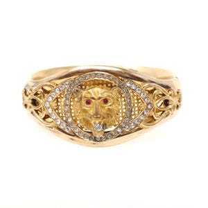 Pode incluir: Uma pulseira dourada com um motivo central de cabeça de leão, olhos de pedra vermelha e uma pedra clara na boca. O leão é rodeado por uma borda circular de strass claros e um intrincado trabalho de filigrana metálica nas laterais.