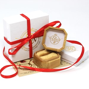 K&ouml;nnte beinhalten: Wei&szlig;e Geschenkboxen mit rotem Band gebunden, eine Box ist ge&ouml;ffnet und zeigt eine goldene Samt-Ringbox. Die Ringbox ist ge&ouml;ffnet und zeigt zwei Ringschlitze. Die Boxen haben goldfarbene Schriftz&uuml;ge und den Text "maejevintage.com".