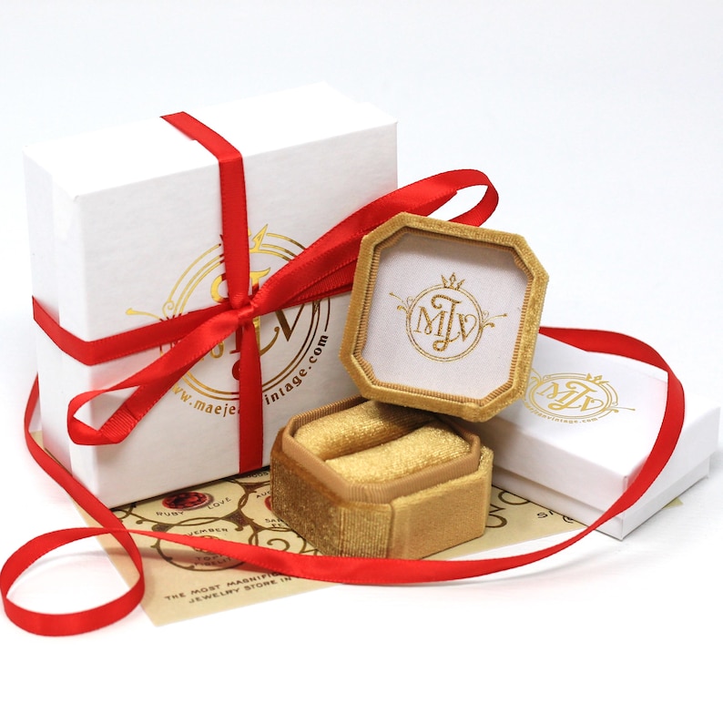 K&ouml;nnte beinhalten: Wei&szlig;e Geschenkboxen mit rotem Band gebunden, eine Box ist ge&ouml;ffnet und zeigt eine goldene Samt-Ringbox. Die Ringbox ist ge&ouml;ffnet und zeigt zwei Ringschlitze. Die Boxen haben goldfarbene Schriftz&uuml;ge und den Text "maejevintage.com".