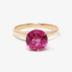 May include: A gold ring featuring a large, round, faceted hot pink gemstone set in a six-prong mounting. The band is smooth and polished, showcasing a classic design with a vibrant, saturated pink center stone that reflects light.