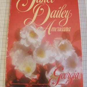 May include: A vintage paperback book titled "Americana" by Janet Dailey. The cover is predominantly red with the title in elegant script and the word "Georgia" in a cursive font. White flowers with yellow centers are featured prominently.
