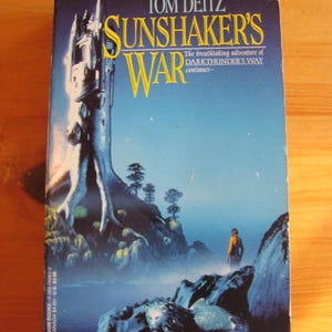 May include: A paperback book titled Sunshaker's War by Tom Deitz. The cover features a tall, dark tower built into a tree, a person standing on a rocky shore, and a lion-like creature in the foreground against a blue gradient background.