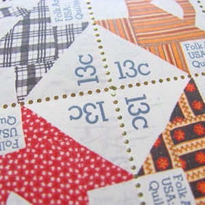 May include: Close-up of vintage postage stamps featuring quilt patterns. The stamps display the text "Folk Art USA: Quilts" and the value "13c". The quilt designs include plaid, striped, and floral patterns in red, black, and orange.