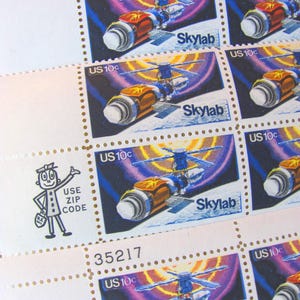 Peut inclure: Feuille de timbres-poste américains vintage représentant la station spatiale Skylab. Chaque timbre présente une illustration bleue, orange et blanche de Skylab, avec le texte "Skylab" et "US 10c". La feuille comprend également un personnage de dessin animé et le texte "Use Zip Code".