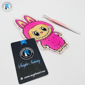 Op de afbeelding: Een roze en wit stripfiguur met een verdrietige uitdrukking, gekleed in een roze konijnenkostuum en een roze pincet. Een zwart label met de tekst "Surglee Industry" is ook aanwezig.