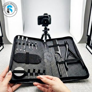 May include: A black zippered case containing various hair styling tools. The case includes combs, clips, and other implements. A camera on a tripod is in the background. The case is held open by two hands.