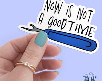 May include: A sticker with the text "Now is Not a good Time" in black lettering. The sticker features a blue seam ripper. The sticker is held by a hand with a gold ring and blue nail polish.
