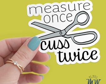 May include: A white sticker with the text "measure once cuss twice" and a grey scissors graphic. The sticker is held against a yellow-green background. The sticker also has the text "MC Woll Designs" in the bottom right corner.