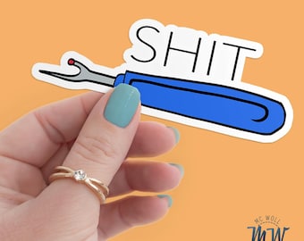 May include: A sticker featuring a blue seam ripper with the word "SHIT" in black capital letters. The sticker is held by a hand with a gold ring and turquoise nail polish against an orange background.